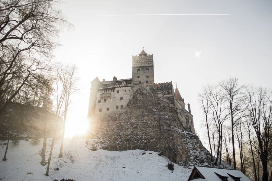 A large castle stands tall on a mountain under a sky with the sun peeking through.