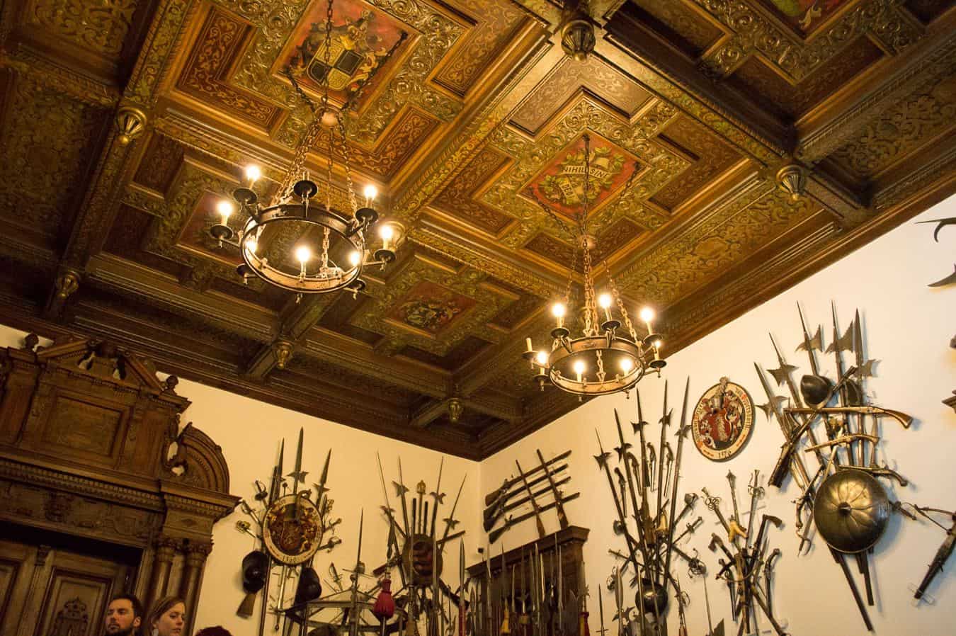 Chandeliers hang from the ceiling in a room that\'s covered in historic weapons.