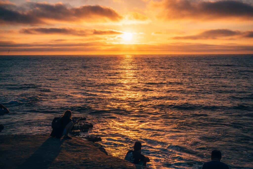 A sunset over a body of water. People sit on rocks watching the sunset.