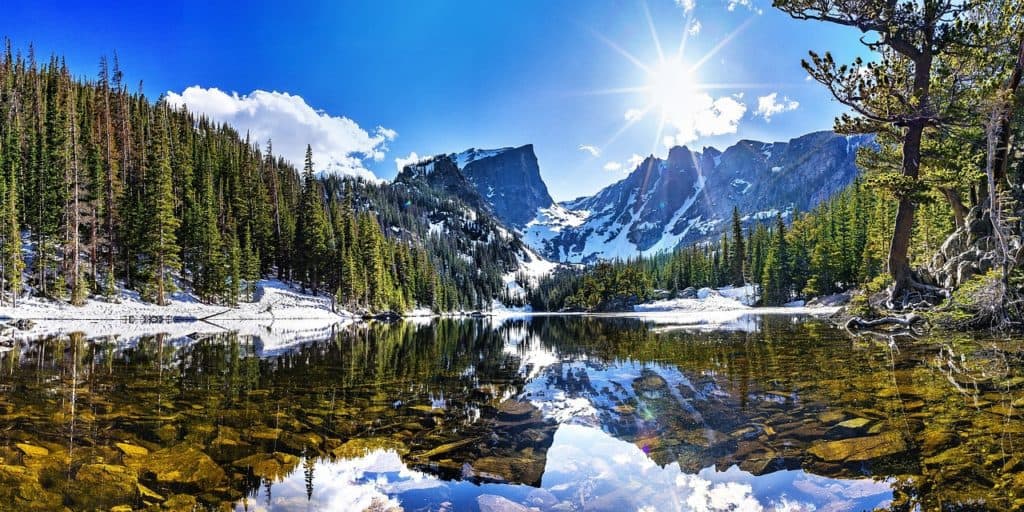 The sun peeks out over a snow-capped mountain and reflects on a calm body of water.