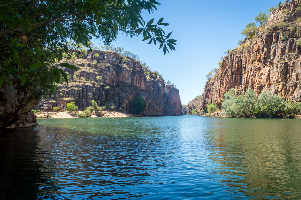 Calm body of water leads into a canyon with tall rock cliffs under a blue sky. 
