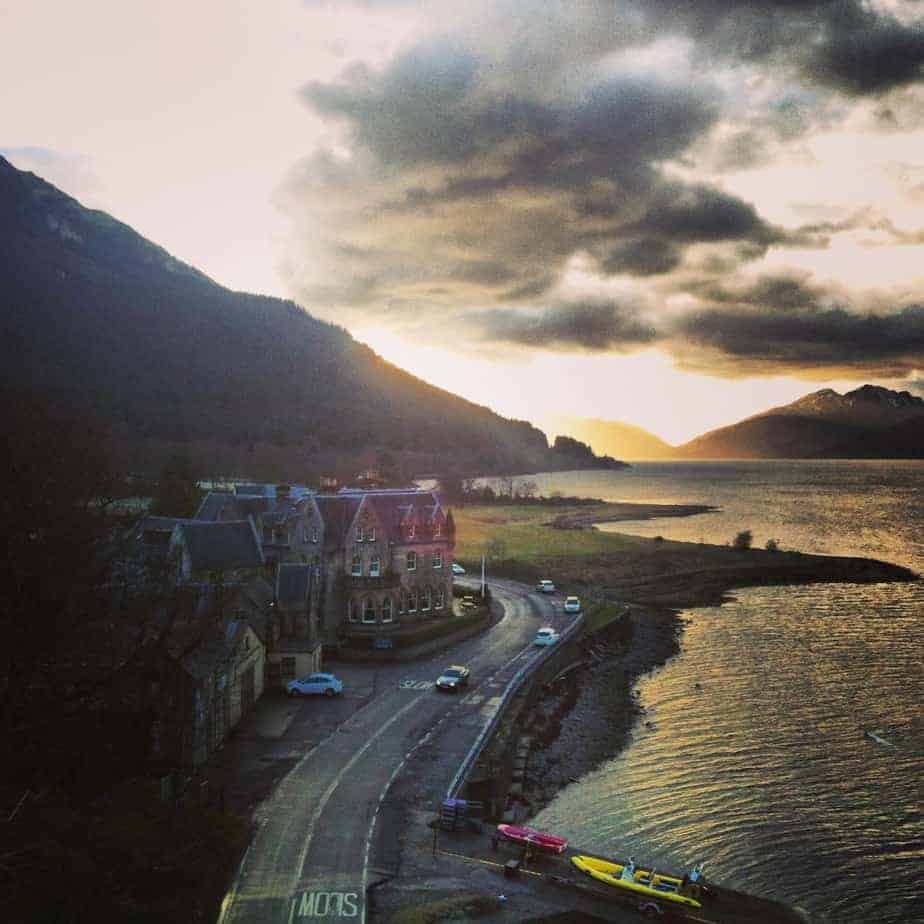 View of a fishing town by the water. A street with cars driving is lined by brick buildings. In the distance, mountains have the sun setting over them.