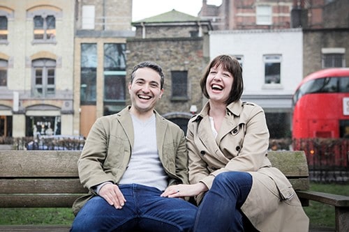 A man and a woman sitting on a bench outdoors, with buildings behind them.