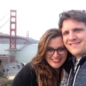 A couple smiling with a big red bridge in San Francisco stretching over water behind them.