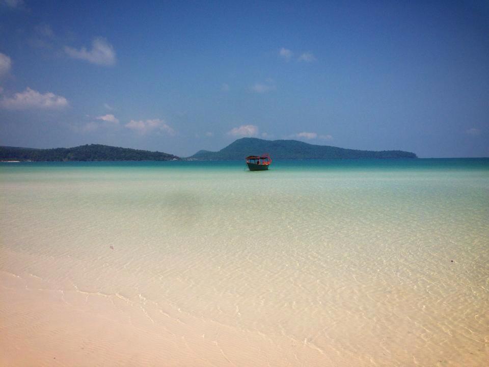 A boat floats on the ocean with sand in the forefront and mountains in the background.