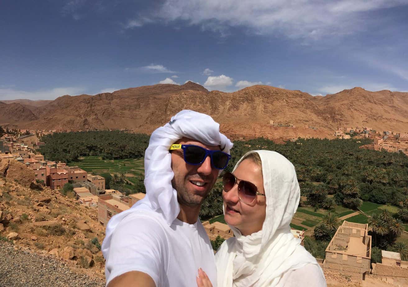 Two people smiling with greenery and rocky cliffs behind them.