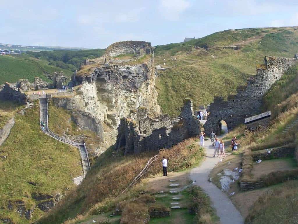 People walk on a gravel path around ruins and mountains.