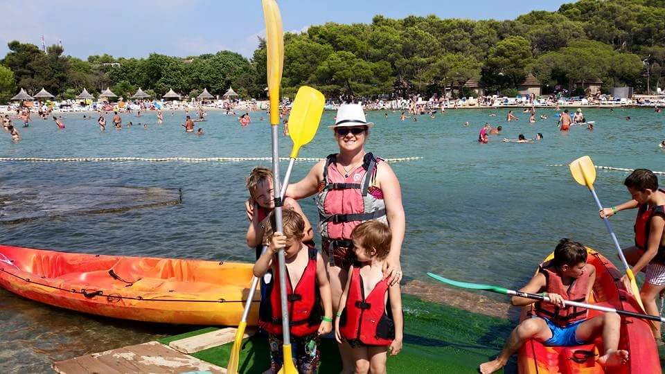 A family stands next to the water all in life jackets, holding yellow paddles.