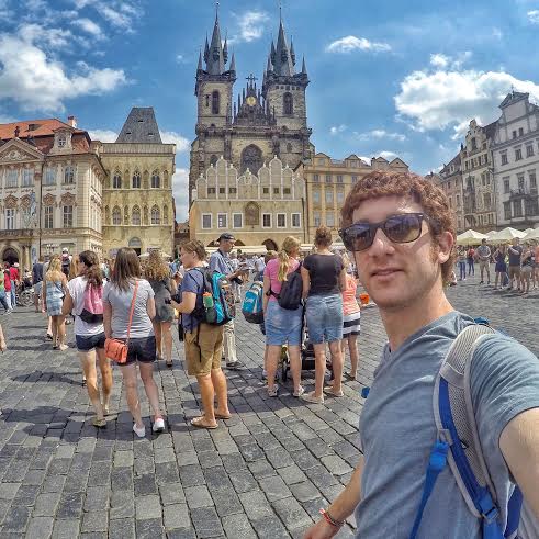 A man smiles while taking a selfie in a grand square filled with people and historic buildings.