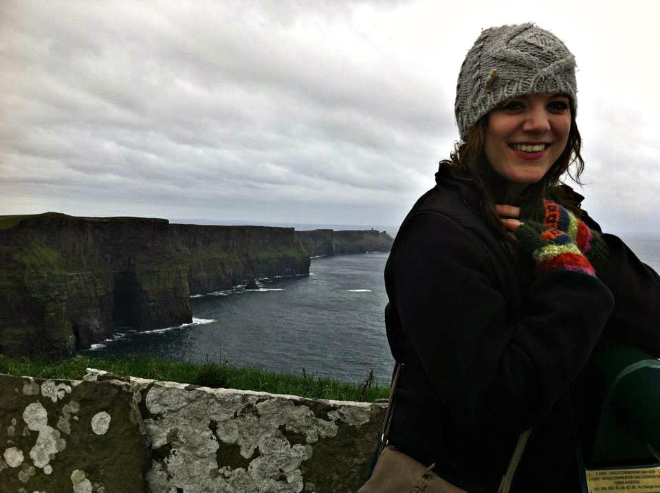 A girl in a grey hat smiles with cliffs and the ocean behind her.