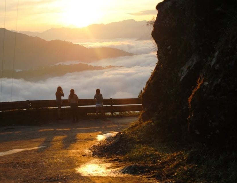 People stand next to a fence looking out at mountain peaks covered in clouds.