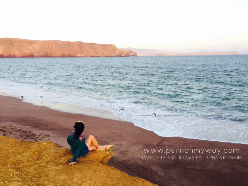 A woman sits in the sand next to the beach.