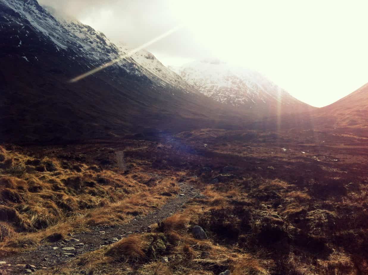 View of a walkway in a field surrounded by snow-topped mountains. The sun shines over the mountains.