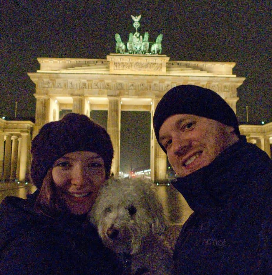 Man and woman smile while holding a dog with an arch structure behind them.