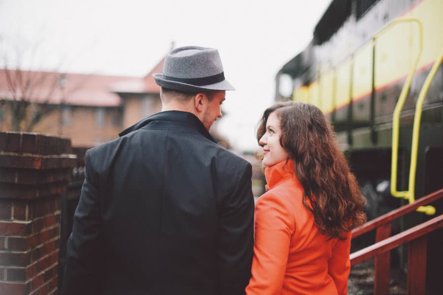 A woman in a red jacket looks at a man with a fedora at a train station.