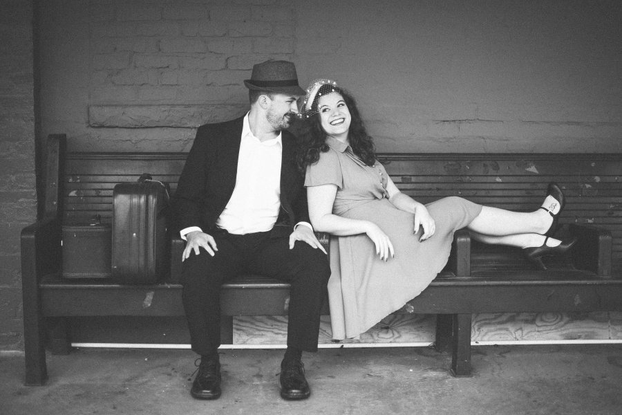 A couple sits on a bench in vintage clothing.