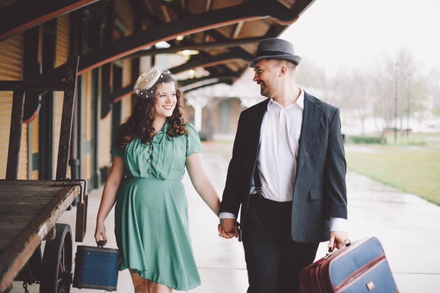 Man and woman in vintage clothing hold hands at a train station.