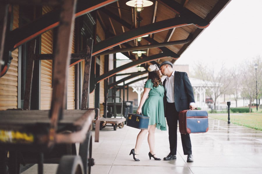A couple kisses at a train station while wearing vintage clothing and holding navy luggage.