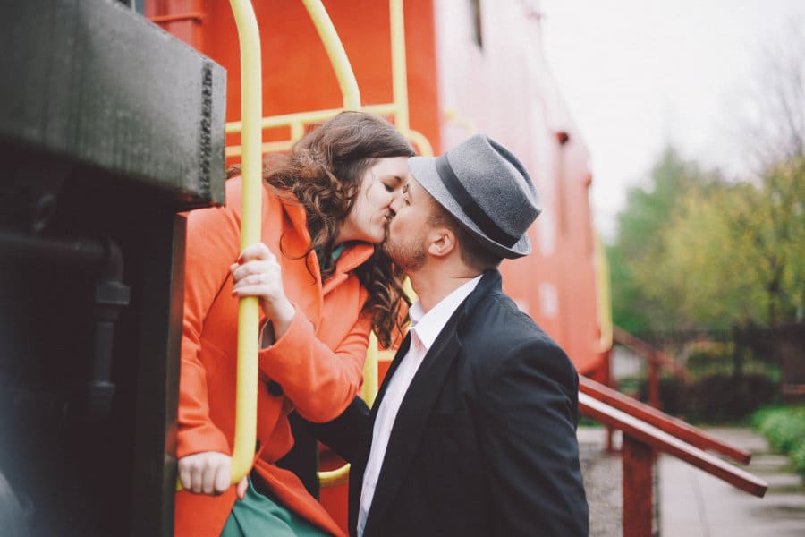 Girl in orange coat leaning out of vintage train to kiss man in suit jacket and hat