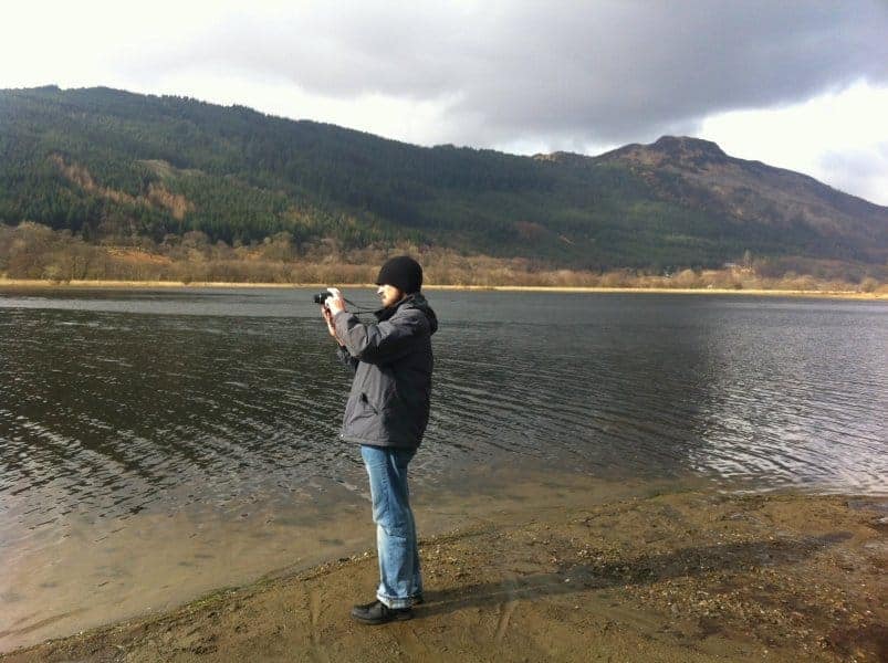 A man with a camera takes a photo over a lake with mountains around.