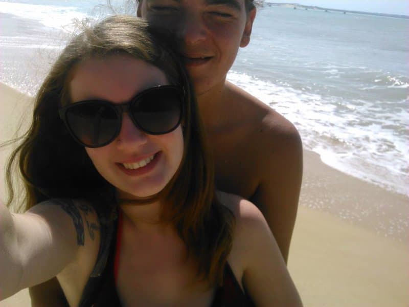 A man and woman smiling on the beach, taking a selfie.