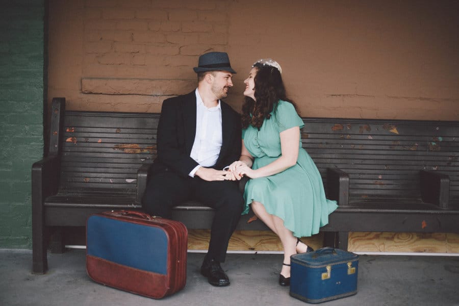 A couple sits on a bench outdoors smiling at one another in vintage clothing.