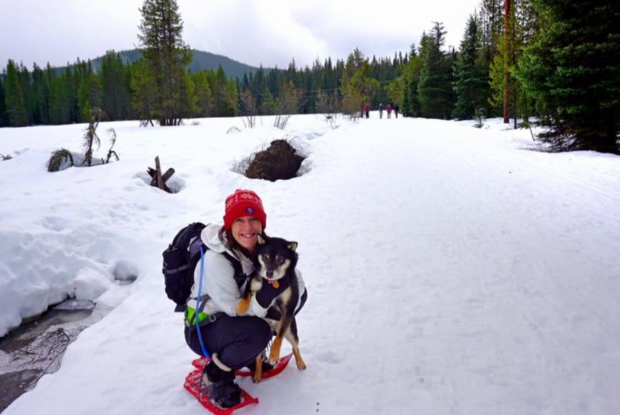 A woman smiles with a dog in the snow surrounded by trees.