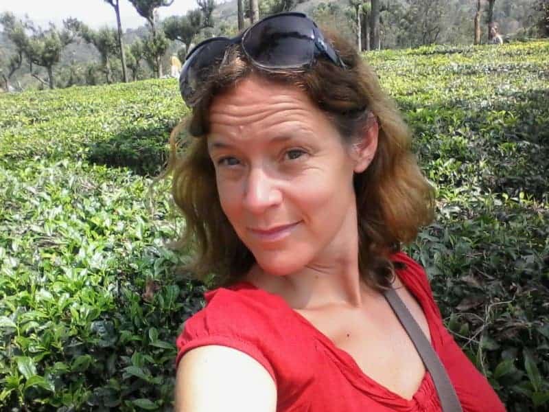 A woman in a red shirt is taking a selfie with green plants behind her.