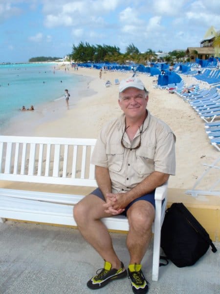 A man sits on a bench smiling with a beach behind him.