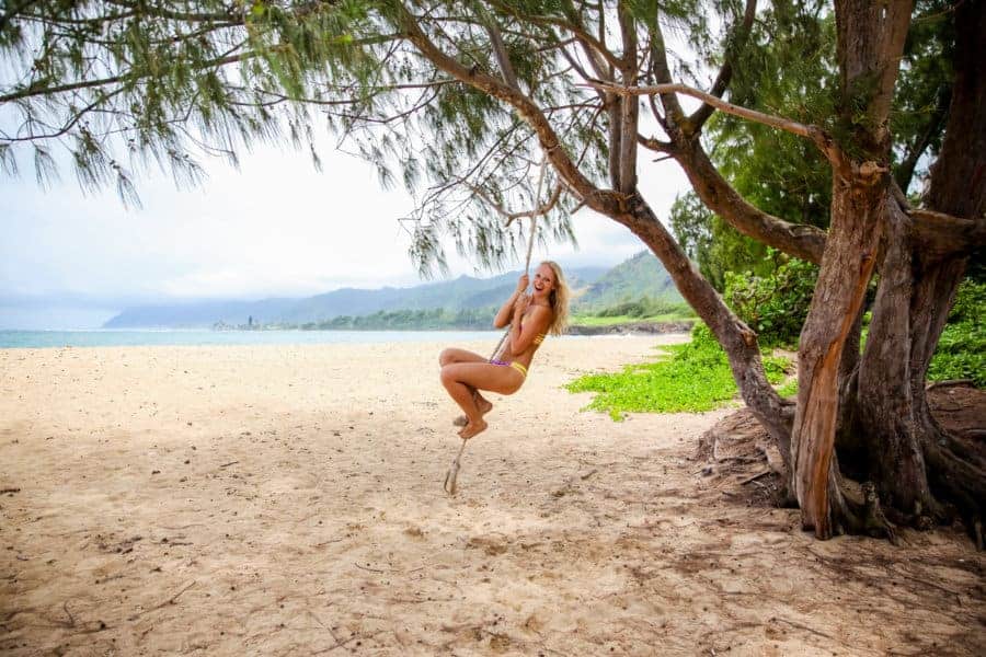 A woman swings on a rope swing hanging off a tree on a beach.