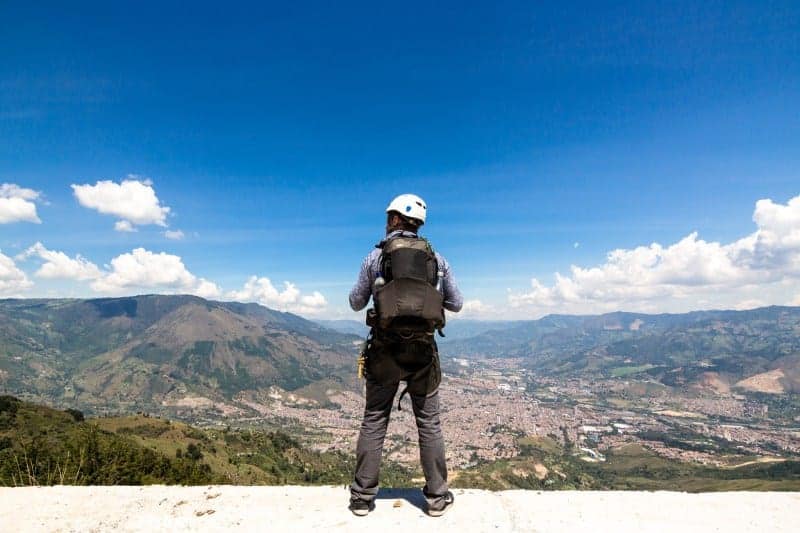 A person stands with a backpack and helmet looking out into a canyon.