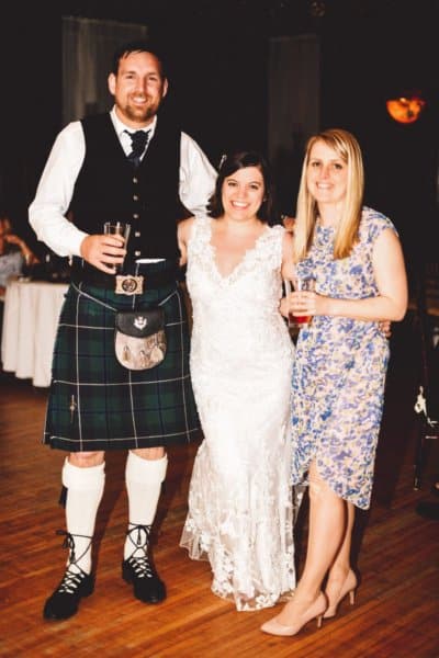 A woman in a wedding dress smiles next to a man in a kilt and a woman in a flower dress.