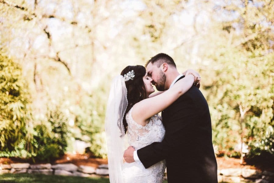 A couple kisses in their wedding outfits with greenery behind them.