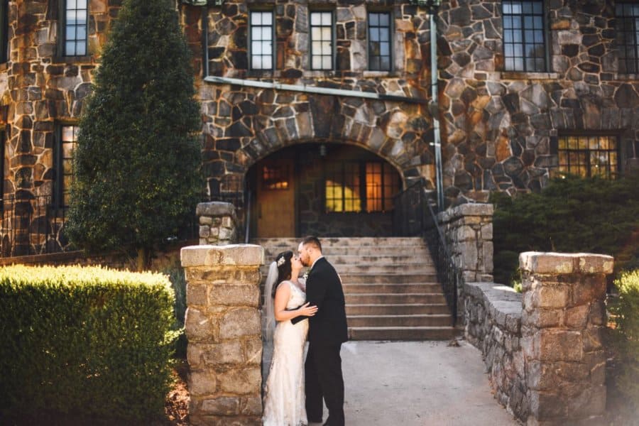 A couple kisses in wedding outfits next to an old brick building.