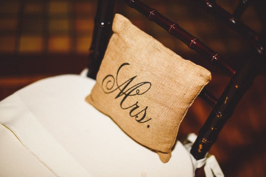 A tan pillow sits on a wooden chair that says \"Mrs.\"