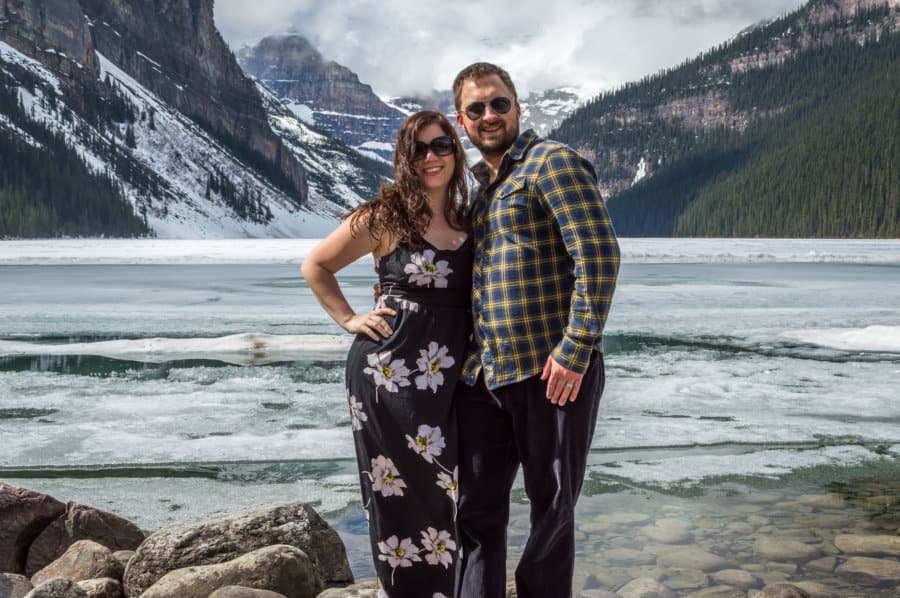A couple smiles in front of a blue lake.