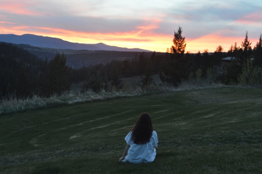 A girl sits in a green landscape looking out at the sun setting.