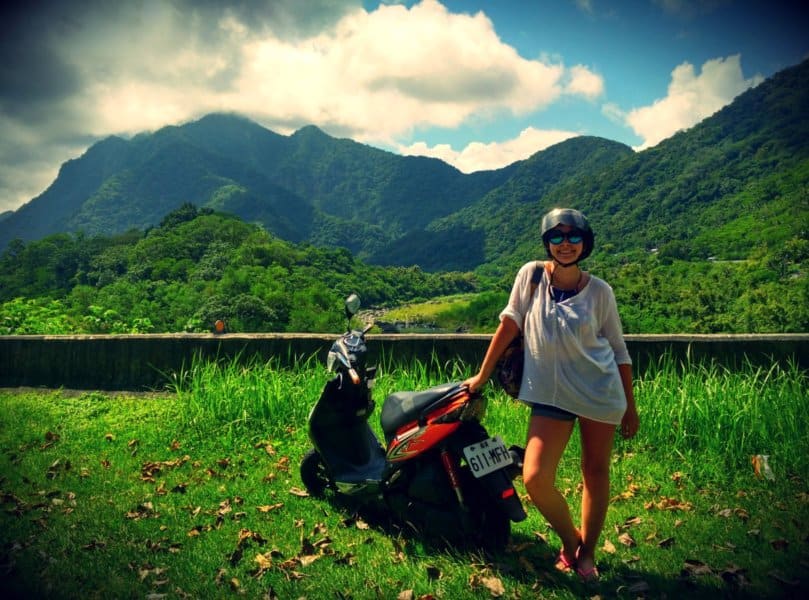 A woman smiles next to a red moped. Green mountains are behind them.