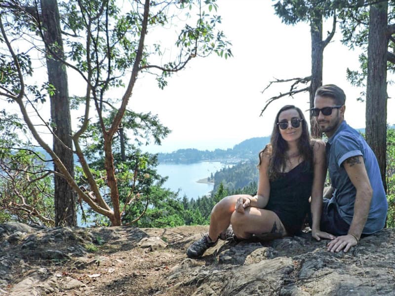 A couple smiles on a rock with a view of the ocean behind them.