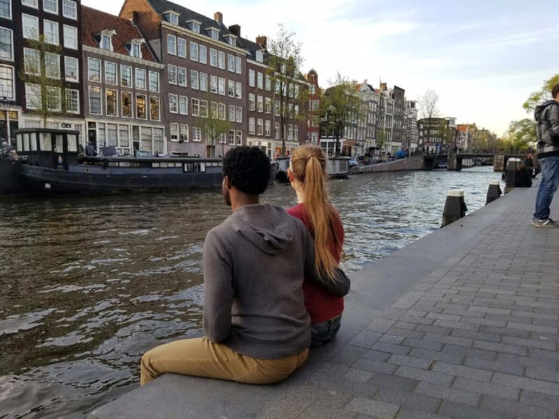 A man and a woman sitting on a sidewalk next to a river.