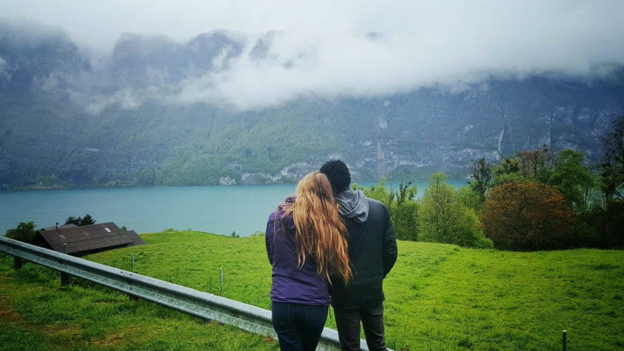A couple stands, leaning against one another, looking out at a view of a lake and mountains.