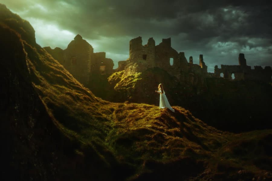 A woman in a white dress stands on a hill with castle ruins behind her.