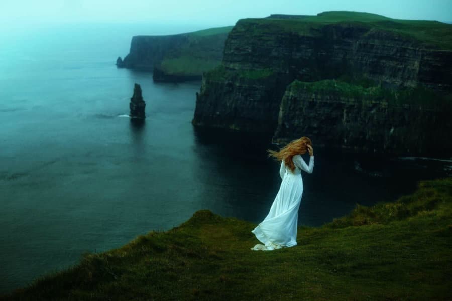 A woman stands in a white dress next to tall cliffs.