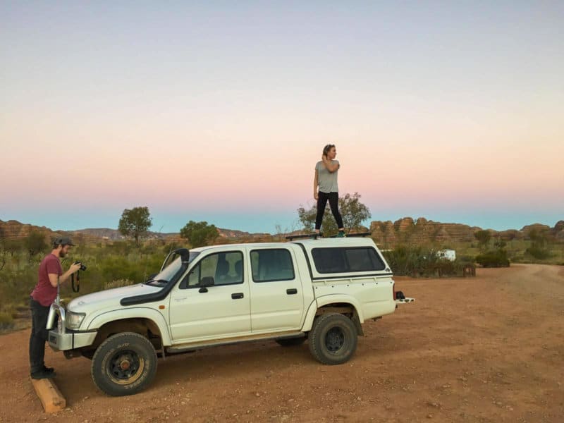 A woman stands on top of a truck while a man is at the front of it with a camera at sunset.
