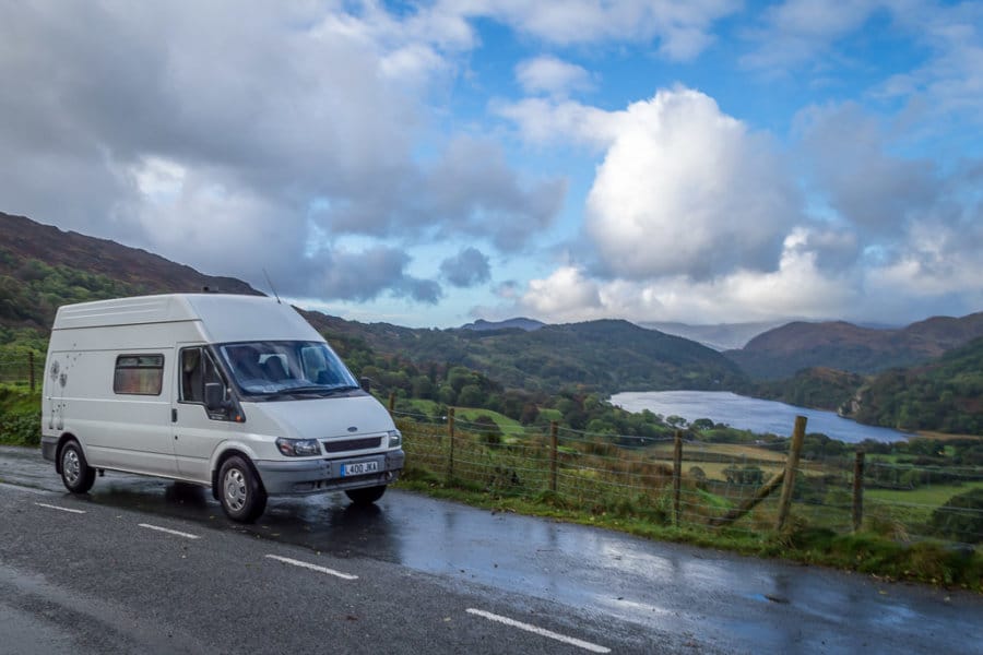 A white van is parked on the side of the road. Mountains and a lake are seen in the background.
