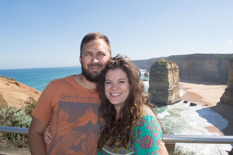 A couple smiles with a beach and cliffs behind them.