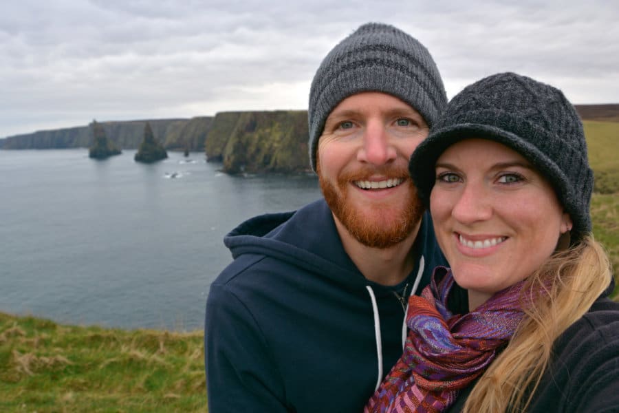 A couple smiles with cliffs near the water behind them.