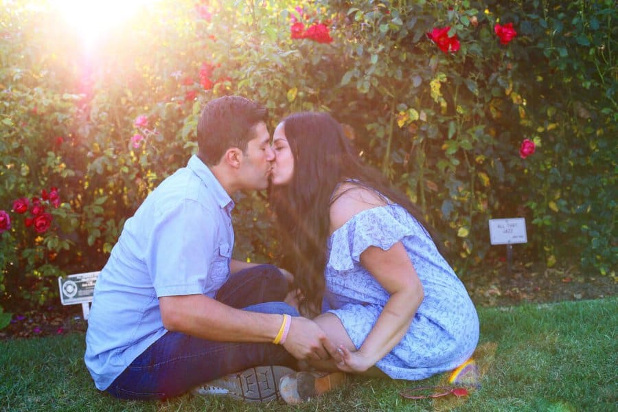 A couple kisses next to a flower bush.