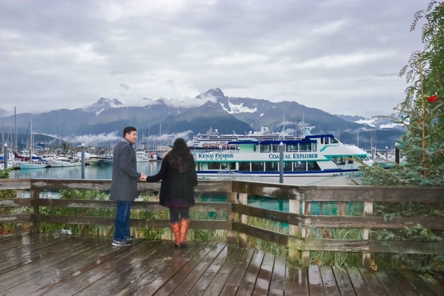 Couple holds hands looking out at a boat floating on blue water. Mountains with snow are in the background.