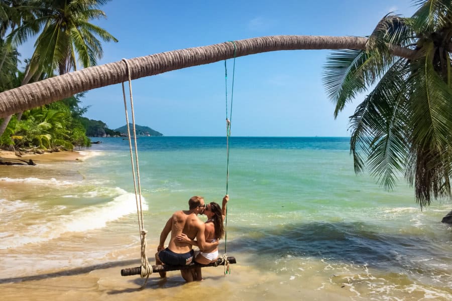 A couple kisses on a swing with a palm tree over the ocean.